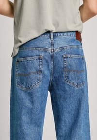Jeans de mezclilla azul con corte de pierna recta, que cuentan con dos bolsillos traseros y una etiqueta de cuero marrón en la cinturilla. Detalle de costura sencilla.