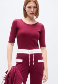 Κολλητό μπορντό τοπ με μανίκια τριών τετάρτων, σε συνδυασμό με τζόγκερ σετ με λευκές λεπτομέρειες και τσέπες· απαλό, ελαστικό ύφασμα.