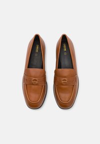 Mocasines de cuero marrón con un acabado suave, que presentan una correa frontal y una puntera redondeada. Suela negra visible abajo. Diseño minimalista.