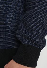 Stickad tröja i mörkblått och svart. Har ett texturerat mönster med ribbade muddar. Mjukt material med en något lös passform.