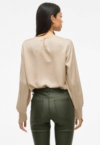 Beige satengbluse med lange ermer, med elastiske mansjetter, sammen med tettsittende olivengrønne bukser med baklommer.