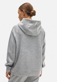 Sudadera gris claro con una textura suave, capucha y mangas raglán. Mostrada desde la parte posterior, acentuando el corte relajado y los detalles de costura.