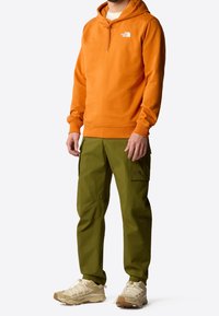 Uomo con una felpa arancione The North Face, pantaloni cargo verde oliva e scarpe da trekking beige in piedi su uno sfondo bianco.