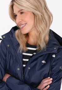 Mornarica vodoodporna jakna z zadrgo spredaj, srebrnim okovjem, z vrvico za nastavljanje kapuce in vezenim logotipom dežnika. Nosi se čez črtasto srajco.