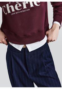 Personne portant un sweat bordeaux avec un texte blanc et un pantalon bleu marine à fines rayures, ajustant la taille avec les deux mains.