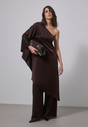 Un mannequin pose en tunique marron foncé à une épaule et en pantalon à jambes larges, tenant une pochette texturée devant un fond de studio uni.