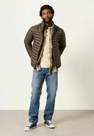 Mann steht mit den Händen hinter dem Rücken, trägt eine grüne Daunenjacke, kariertes Hemd, beiges Henley-Shirt, blaue Jeans und navyblaue Sneakers auf grauem Boden.