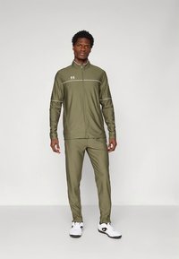 Olivno zelena trenerka z zip-up jakno z belimi poudarki, združena z ujemajočimi hlačami in belimi športnimi čevlji.