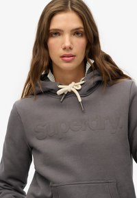 Un sweat à capuche gris avec un logo "Superdry" texturé, une capuche avec cordon de serrage et un col doublé. Il possède une poche kangourou à l'avant et une texture de tissu douce.