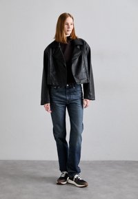 Veste en cuir noire coupée sur un top sombre, associée à un jean en denim bleu et des baskets noires avec des accents blancs, se tenant sur un fond neutre.