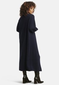 Donna con capelli ricci che indossa un lungo cappotto blu navy con trama e stivali neri con tacco, in piedi con il corpo parzialmente girato di schiena.