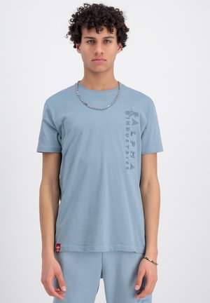 Helles blaues Baumwoll-T-Shirt mit kurzen Ärmeln, Rundhalsausschnitt und einem vertikalen "Alpha Industries"-Logo. Verziert mit einem roten Logotag am Saum.