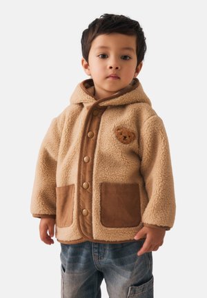 Giacca in pile beige con accenti marroni, bottoni a pressione e due tasche frontali. Presenta un'applicazione di orso sul petto.