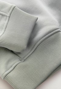 Jasnoszary mankiet bluzy z charakterystycznym prążkowanym wykończeniem, gładkim materiałem i detalami w postaci ściegów. Skoncentruj się na miękkim materiale i funkcjonalnym designie.