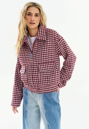 Blondynka w czerwono-białej kurtce w kratę pepitkę z niebieskimi detalami zamka błyskawicznego oraz jasnoniebieskich dżinsach dwukolorowych na białym tle.