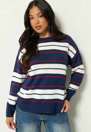 Jeune femme aux longs cheveux foncés portant un pull bleu marine avec des rayures horizontales blanches et rouges et un jean bleu sur un fond uni.