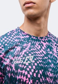 Κοντινό πλάνο ενός ατόμου που φοράει ένα ροζ και σκουρομπλε πουκάμισο με μοτίβο από αφηρημένα ψηφιδωτά τετράγωνα και λευκό λογότυπο κειμένου στο στήθος.