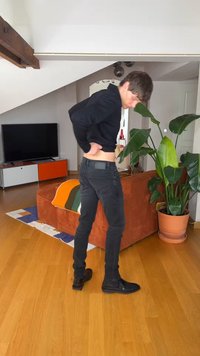 Noor mees mustas särgis ja teksades kohandamas pükse elutoas, kus on oranž diivan, värvikas tekk, suur taim ja telerikapp.