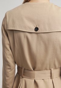 Trench beige con un cuello estructurado, cintura ceñida con cinturón y un detalle de botón negro brillante en la parte posterior. Textura de tela suave con costuras a medida.