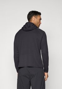 Svart hettegenser med et teksturert mønster, lange ermer og en lett formsydd silhuett. Har en buet nederkant og raglan-ermer.