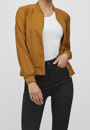 Femme portant une veste aviateur moutarde sur un haut blanc et un pantalon taille haute noir, une main dans la poche et l'autre détendue le long du corps.