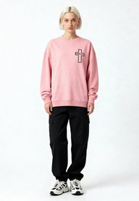 Jeune personne debout, portant un sweat-shirt rose avec une croix noire et le texte "Karma Kills", un pantalon cargo noir et des baskets blanches.