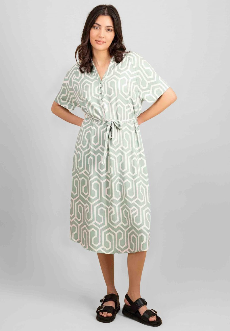 Rochie verde deschis cu un model geometric, mâneci scurte, închidere cu butoane și talie legată, purtată cu sandale negre.