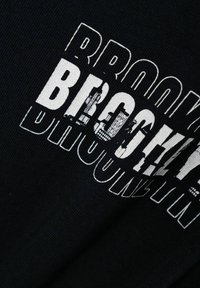 Camiseta de tela negra con un audaz texto gráfico en blanco que dice "BROOKLYN", con un acabado desgastado y tamaños de fuente variados para resaltar.