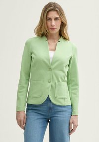 Blazer vert clair avec une texture côtelée, fermeture à un bouton et deux poches avant. Associé à un haut blanc et un jean bleu.