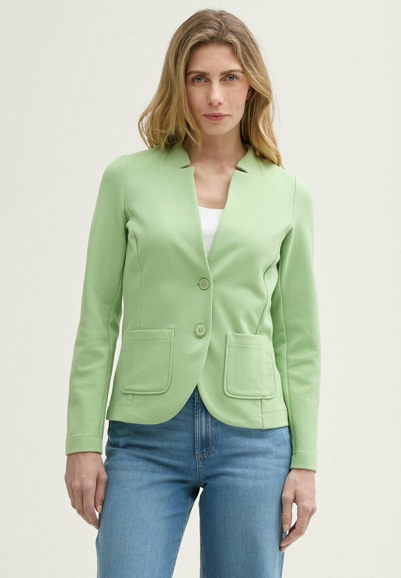 Blazer vert clair avec une texture côtelée, fermeture à un bouton et deux poches avant. Associé à un haut blanc et un jean bleu.