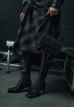 Czarne skórzane kozaki za kolano z kwadratowymi noskami i blokowymi obcasami, zestawione z kratkowaną spódnicą i plecioną czarną torebką.