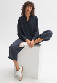 Marineblaue Bluse mit langen Ärmeln und lockerer Passform, kombiniert mit strukturierten dunklen Hosen. Weiße Sneakers mit hellbraunen Sohlen runden das Outfit ab.