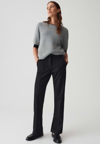 Vrouw in een grijze korte, fuzzy trui met korte mouwen, een zwart shirt met lange mouwen eronder, zwarte op maat gemaakte broeken en zwarte instappers, staande met haar handen in de zakken.
