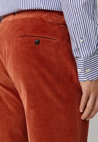 Rust corduroy trousers featuring vertical ribbing, a single back pocket with a black button, and a smooth texture. Paired with a striped shirt.
