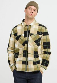 Veste en polaire de couleur beige, noire et blanche à carreaux avec des accents verts, présentant une fermeture éclair à l'avant et deux poches poitrine. Col et poignets côtelés.