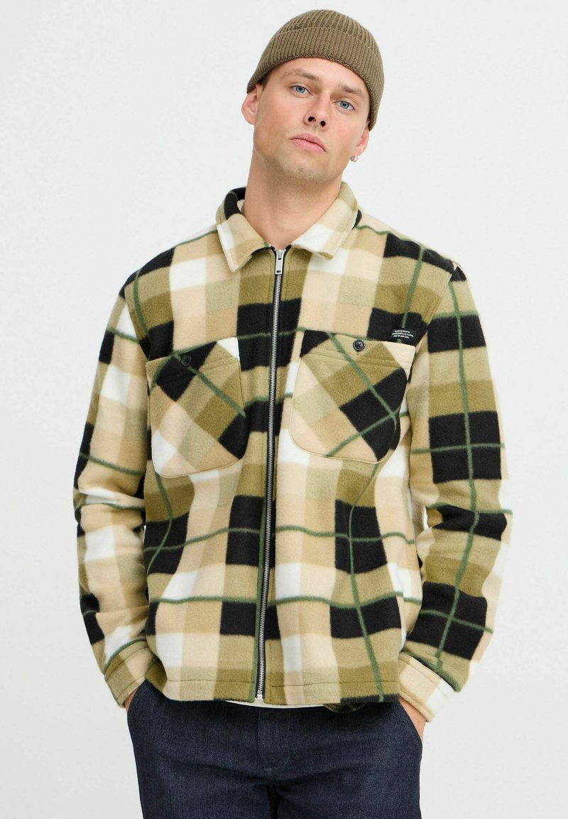 Veste en polaire de couleur beige, noire et blanche à carreaux avec des accents verts, présentant une fermeture éclair à l'avant et deux poches poitrine. Col et poignets côtelés.