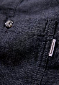 Mörkblå vävt tyg med ett fiskbensmönster, som har en rund knapp och en etikett som visar "LINDBERGH" i vitt. Subtila texturdetaljer.