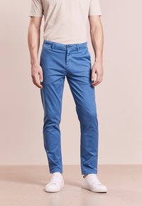 Modré bavlněné chinos kalhoty s úzkým střihem, s knoflíkovým zapínáním a poutky na pásek, kombinované s bílými teniskami na neutrálním pozadí.