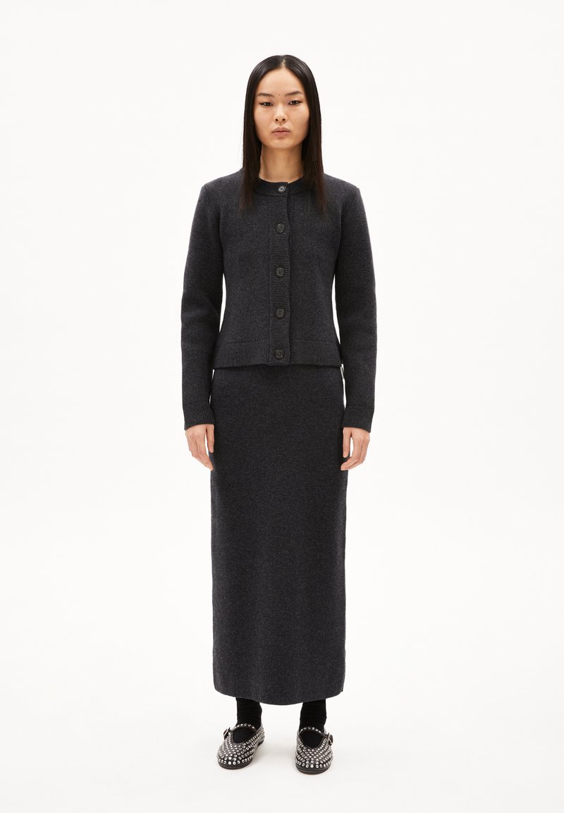 Frau steht in einem dunkelgrauen, geknöpften Cardigan, passendem langen Rock, schwarzen Socken und besetzten schwarzen Slippern vor einem weißen Hintergrund.