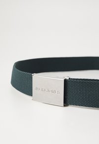 Elastický pásek v tmavě zelené barvě, s kovovou sponou v stříbrné barvě s vyrytým nápisem "JACK & JONES." Texturovaná tkanina s hladkým povrchem.