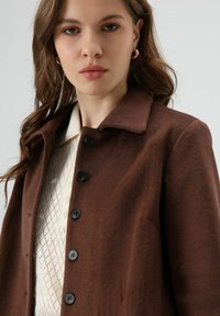 Bruine jas van zacht pluche stof met een kraag, voorzien van zwarte knopen en een getextureerde witte trui eronder. Glad oppervlak, op maat gemaakt ontwerp.