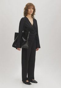 Schwarzes Wickeltop mit langen Ärmeln, kombiniert mit weiten schwarzen Hosen und spitzen schwarzen Ballerinas. Eine große, glänzende schwarze Handtasche rundet das Outfit ab.