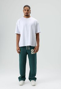 Witte oversized katoenen T-shirt gecombineerd met donkergroene joggingbroek met een beige genaaide "22". Model draagt witte sneakers.