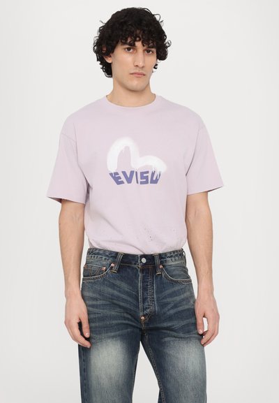 T-shirt en coton violet clair avec un logo graphique "EVISU" blanc et bleu, associé à un jean en denim bleu délavé avec des accessoires en cuivre.