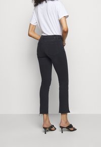 Mujer con vaqueros negros recortados con dobladillos sin rematar, camiseta blanca de manga corta y sandalias negras con tacón, de pie frente a un fondo claro y sencillo.