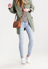 Giacca leggera verde oliva, top con stampa leopardata, jeans azzurri strappati e sneakers bianche. Borsa a tracolla marrone con cinturino alla spalla.