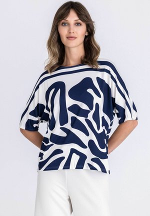 Vrouw met golvend haar tot op de schouders, gekleed in een witte en marineblauwe, abstracte, kortärmelige top en witte broek, staand met haar handen achter haar rug.