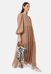 Robe en chiffon rose, longue et à volants, avec des manches bouffantes. Les accessoires comprennent un petit sac à main beige et une écharpe à motifs floraux. Talons carrés.