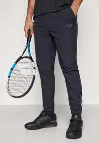 Melni sporta bikses, kas izgatavotas no viegla materiāla, ar šauru piegriezumu, ar sānu kabatām un baltu svītru pie potītes. Rokā tur tenisista rakete.