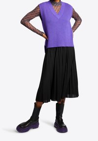 Lila geripptes ärmelloses Top, schwarzer plissierter Rock und klobige schwarze Stiefel mit lila Sohlen, das Top mit einem auffälligen geometrischen Muster.